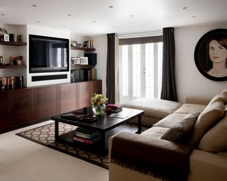 White basement living room, spot lighting, beige sofa, black coffee table, biult in plasma television, book shelves, built in wooden units, patio doors, oval framed oil painting.