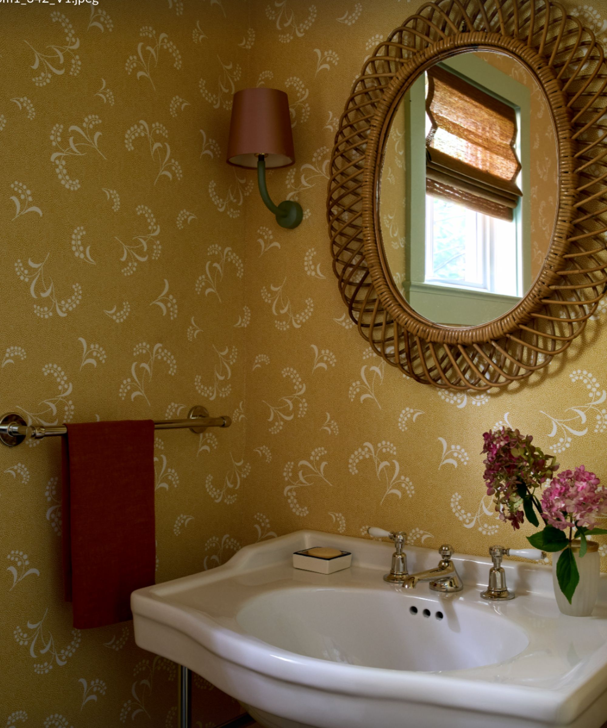 Charming powder room with yellow floral wallpaper, a white pedestal sink, and a round rattan mirror. A small wall sconce and pink flowers add a cozy, vintage feel.