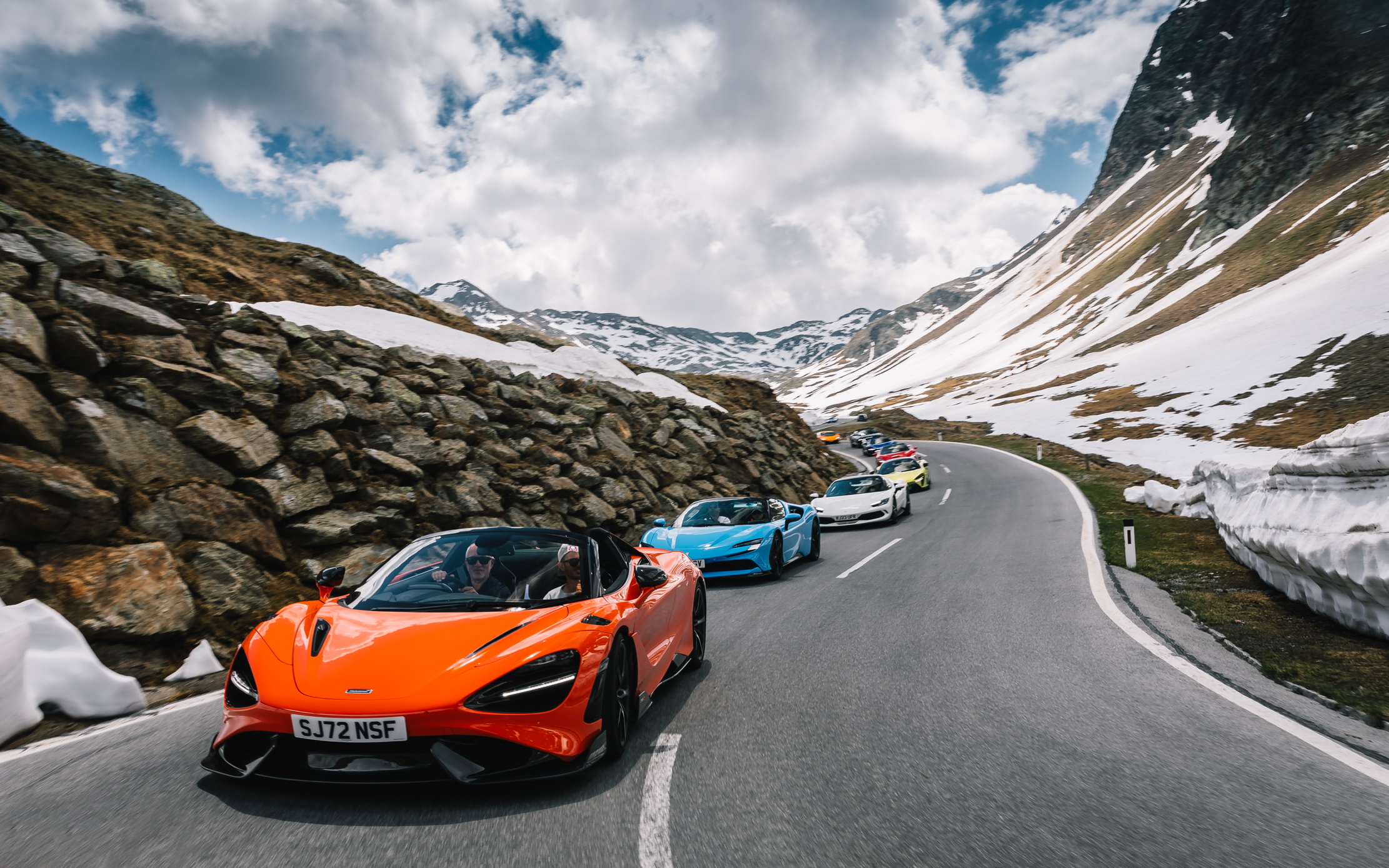 A line of supercars, including a McLaren, A ferrari, another ferrari and a porsche weave their way through the Italian Dolomites