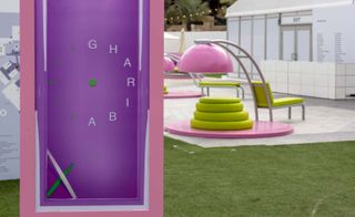 Outside daytime image, lawn area, grey patio space, white building with windows and exit door, close up of a tall purple and pink clock style exhibit, seating incorporating a ladder pink, green and grey art work