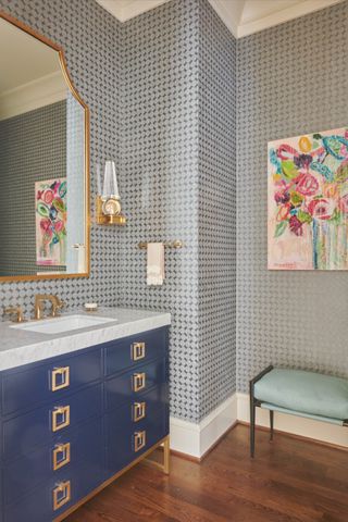 bathroom with grey patterned wallpaper, dark wooden floor, blue vanity unit, artwork, mirror, stool