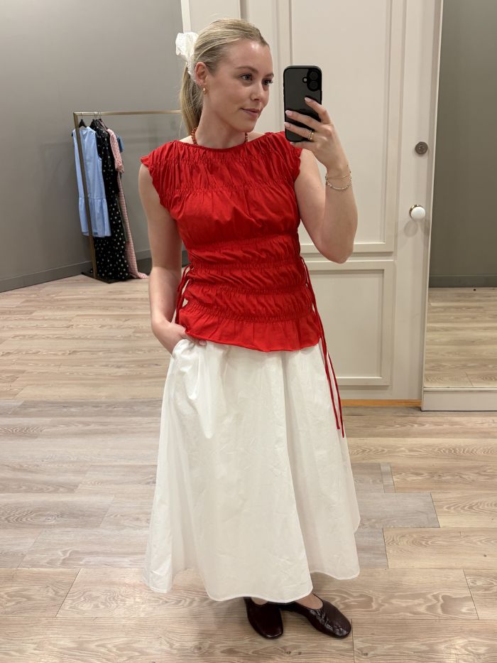 Woman wears red ruched top, white skirt and burgundy ballet flats