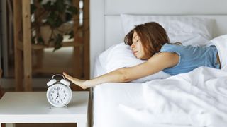 A woman reaches out of bed to press the snooze button on her alarm clock