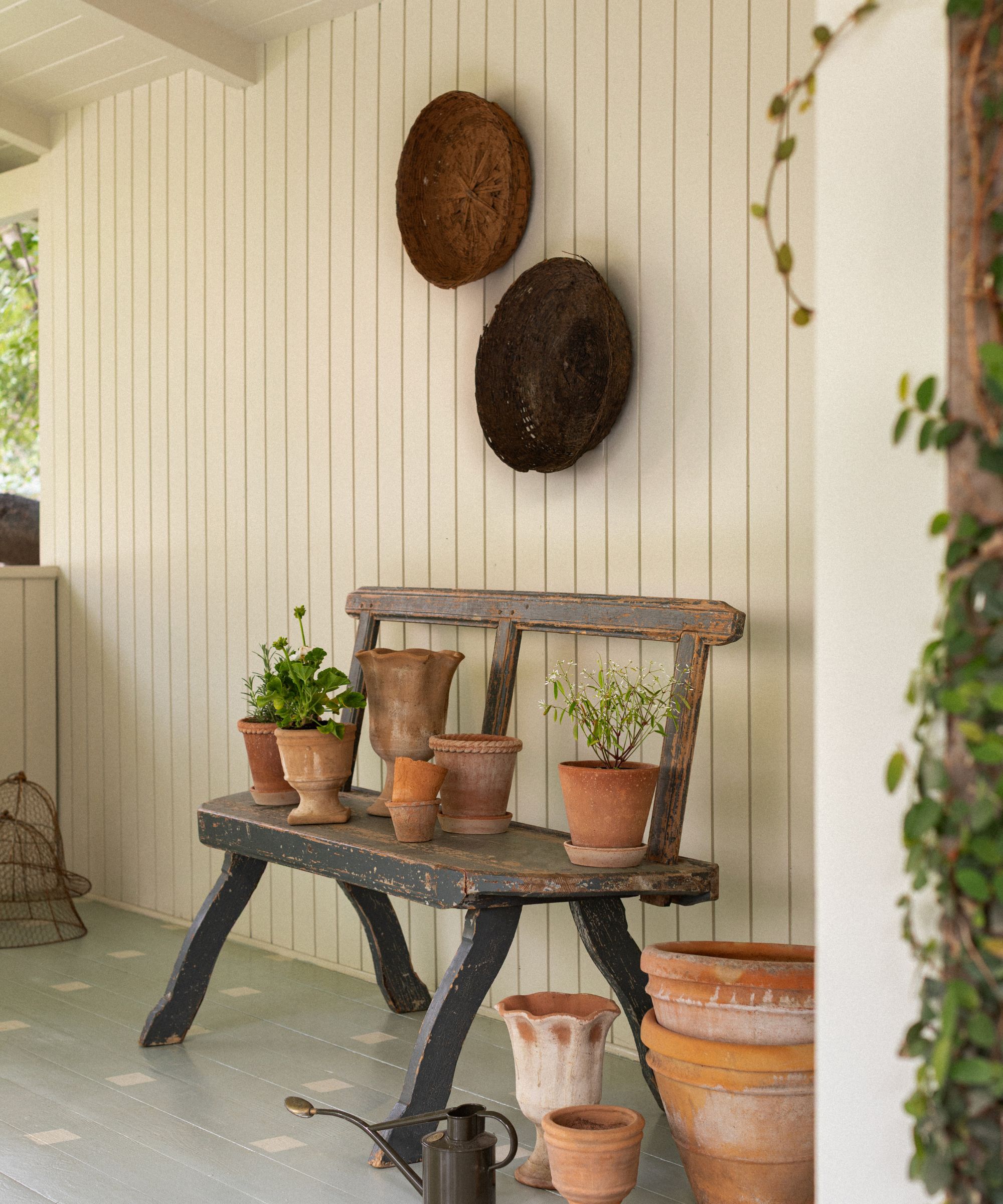 An outdoor porch with creamy white wood-clad walls, a wooden bench with terracotta plant pots, and two baskets displayed on the wall.
