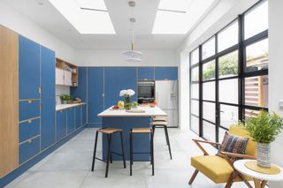 Wide shot of kitchen with grey large-format floor tiles, blue and pink Formica plywood units, white worktop and Crittall-style doors to garden