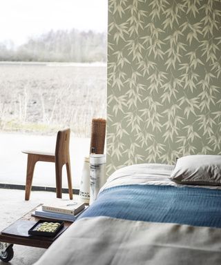 A green-grey wallpaper with bamboo leaf print motif in bedroom with grey duvet and wooden chairs