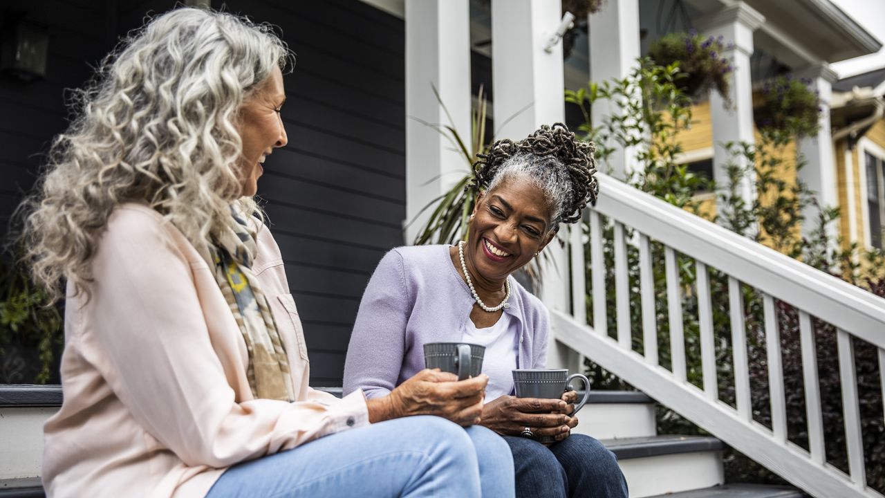 Two older women laugh as they sit on porch steps and drink coffee.