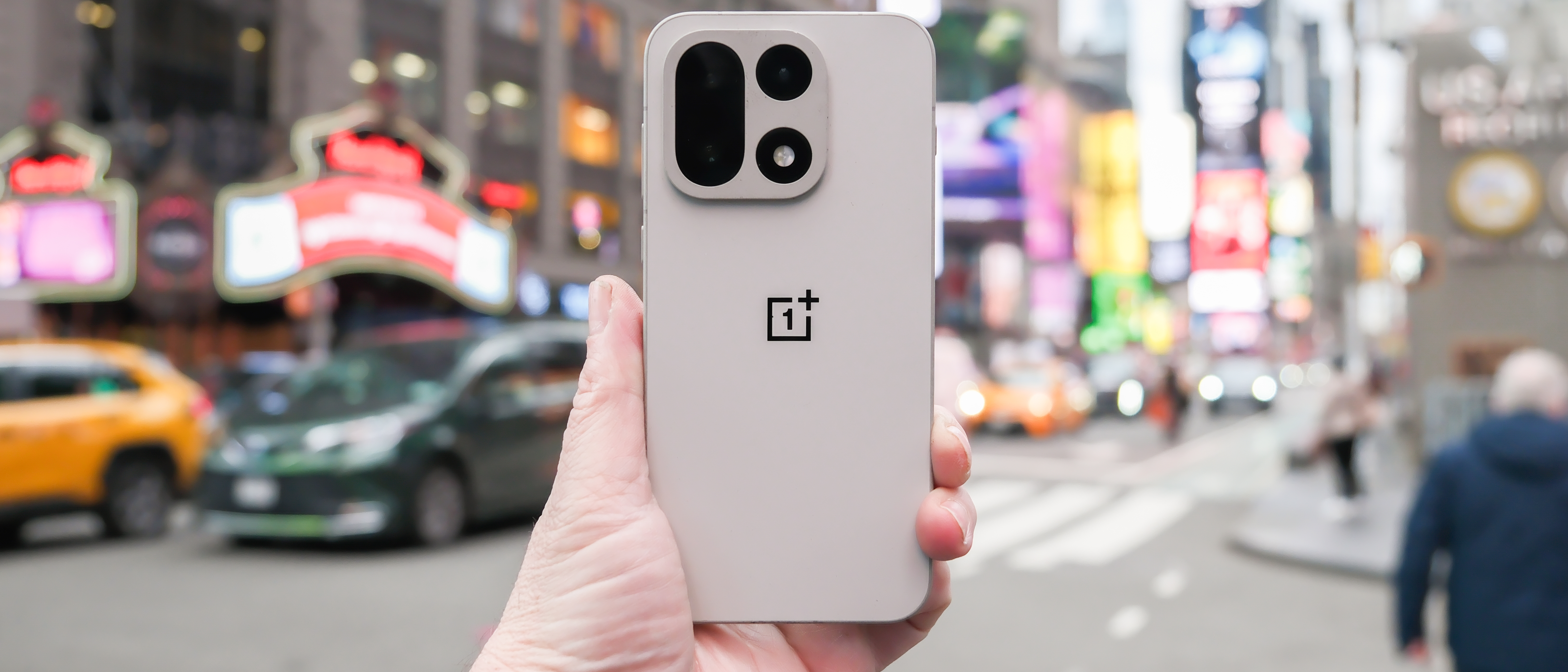 OnePlus 15 in Sand Storm color held in hand with cameras and back showing in the middle of a Times Square New York City intersection across from the Hard Rock Hotel. The background is blurred with cars and people moving past