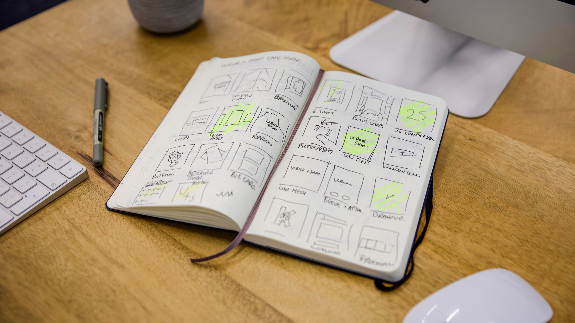An open notebook on a wooden desk shows design sketches and diagrams highlighted in yellow marker next to a keyboard, mouse, and monitor.