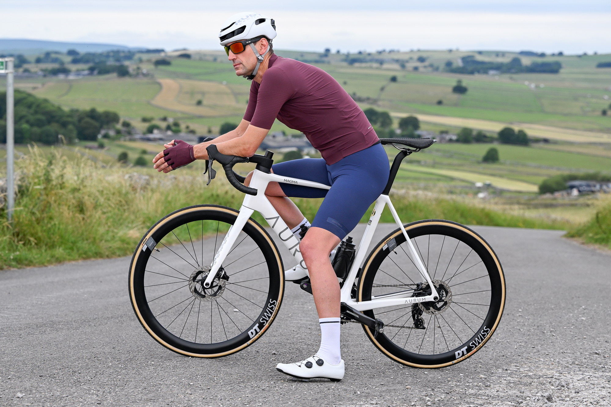 Man sitting on a white bike wearing a purple jersey, purple gloves, blue shorts and a white helmet