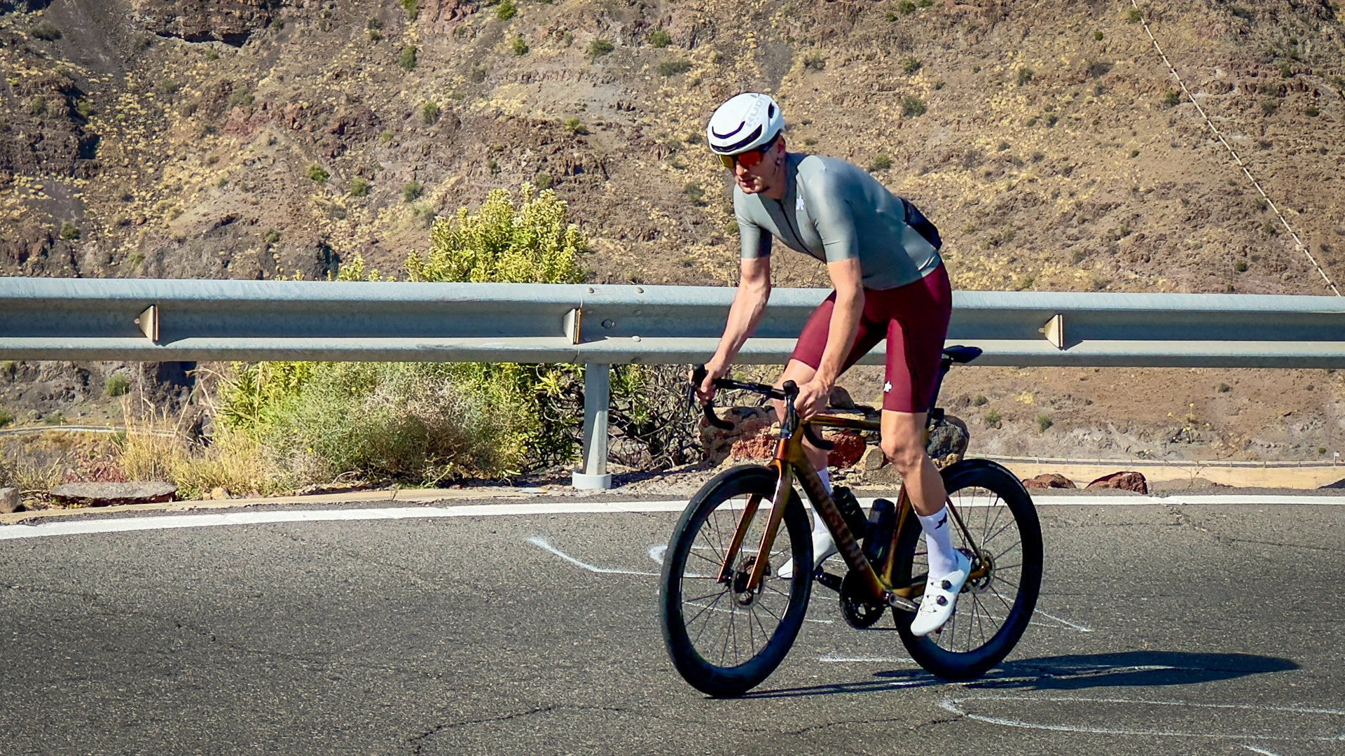 Man wearing a green jersey, red shorts and white helmet riding a road bike up a hill out of the saddle