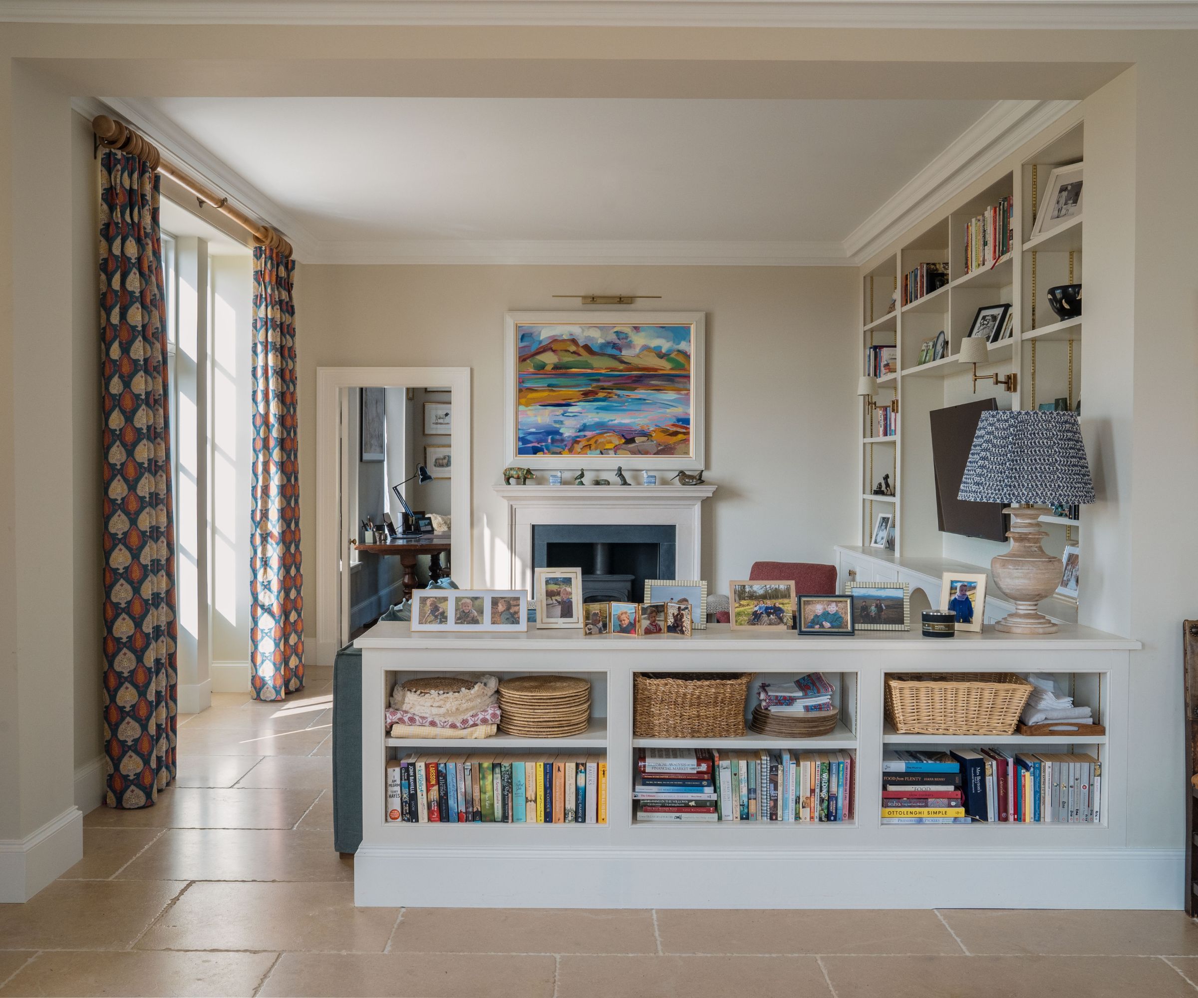 Living room with tiled floor, cream walls, French doors with floral drapes, a built-in storage unit with books, trinkets, and a TV at the centre, and a storage island