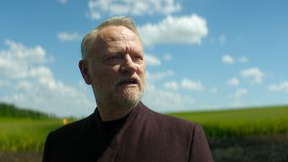 A bearded man in a green field under a cloudy blue sky