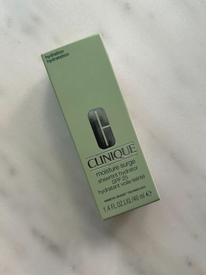 The Clinique Moisture Surge Sheertint Hydrator SPF 25 in its green box on a marble table