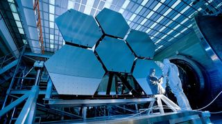 A couple of people wearing white outfits stand in front of the large mirrors of the James Webb Space Telescope