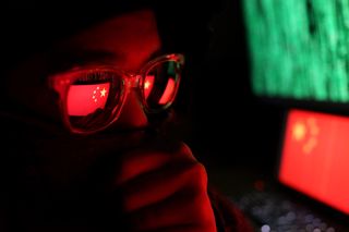 A hacker wearing glasses in a dark room with the Chinese flag shown in the background on a computer display