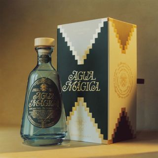 A bottle of Agua Mágica mezcal next to its black, white and gold box pictured against a brown background