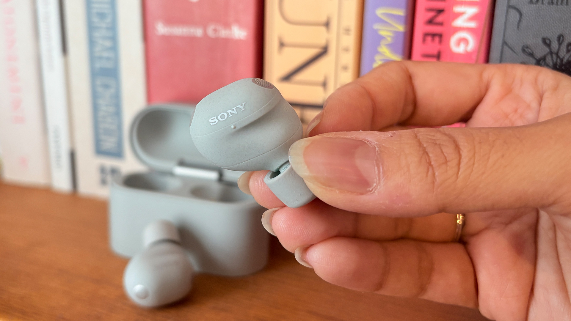 Sony WF-1000XM6 wireless earbuds, one held in hand in front of case and bookshelf