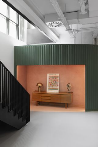 A green structure housing pink workspace with a wood side table, brass lamp and artwork. Located inside Black Kite office, London