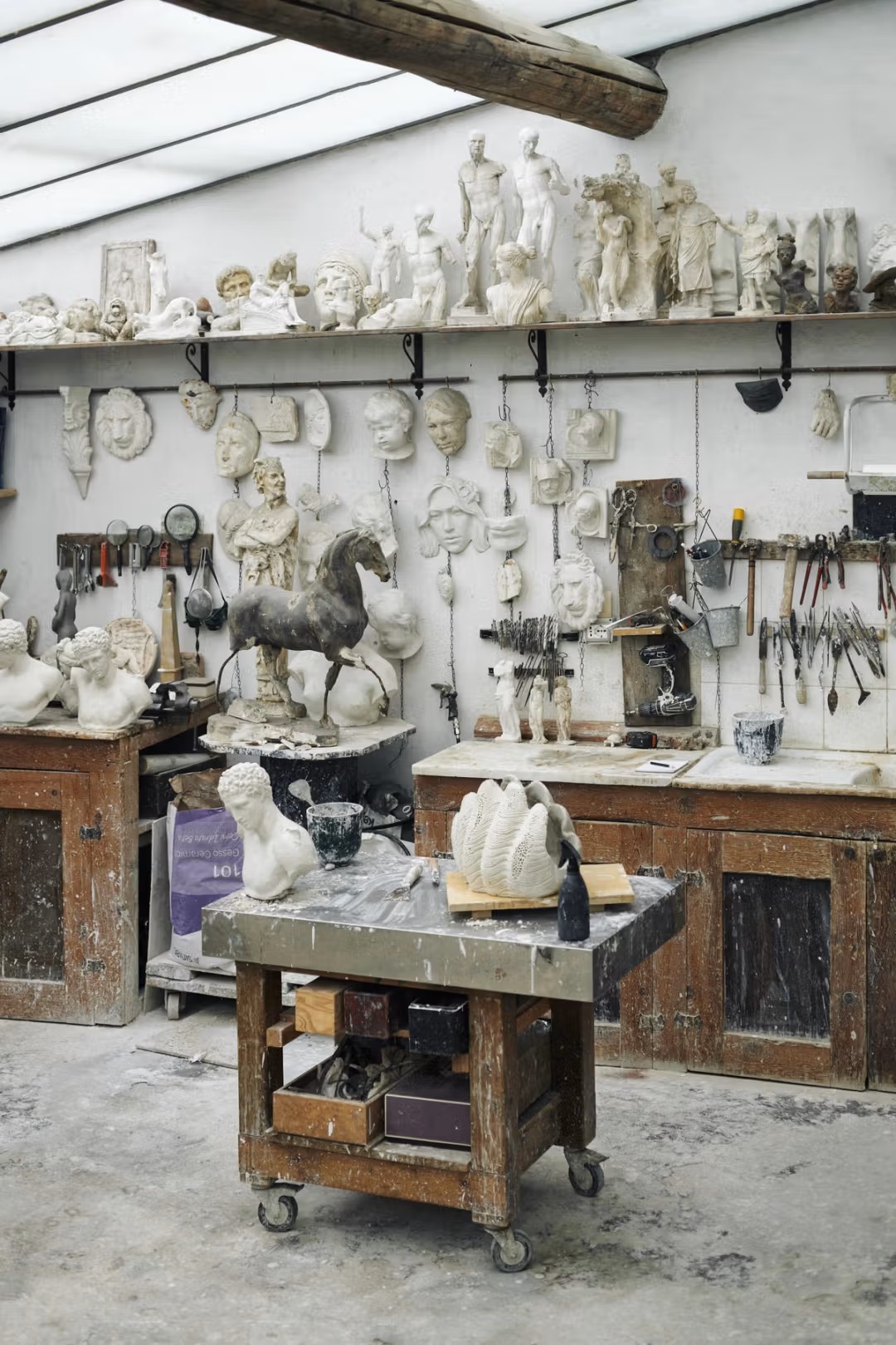 A marble sculpture studio filled with masks, statuettes, and other decorative objects still being finalized, decorated with industrial cement flooring and wooden shelving units.