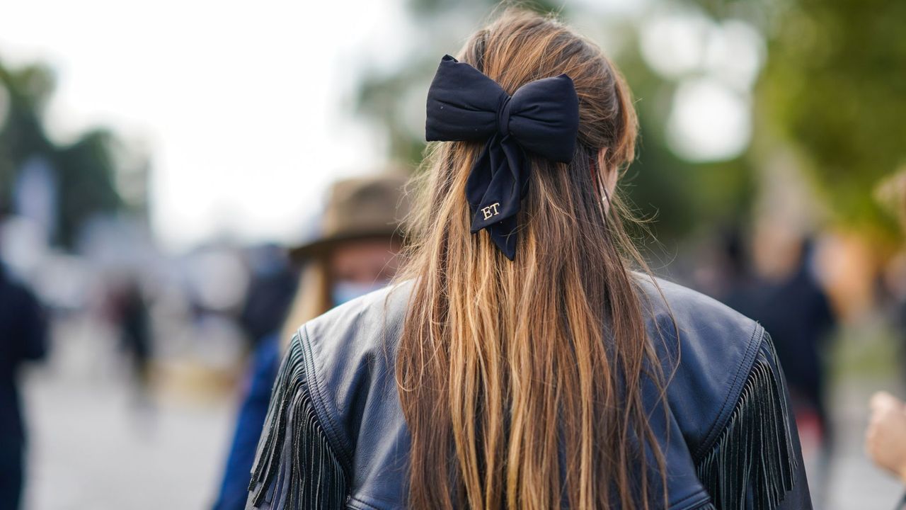 Argan oil for hair: A woman with long brown hair and blonde balayage wearing a black bow in her hair and a black leather jacket