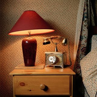 Brown bedside cabinet with red table lamp and a telphone photographed against a wall with patterned wall paper.