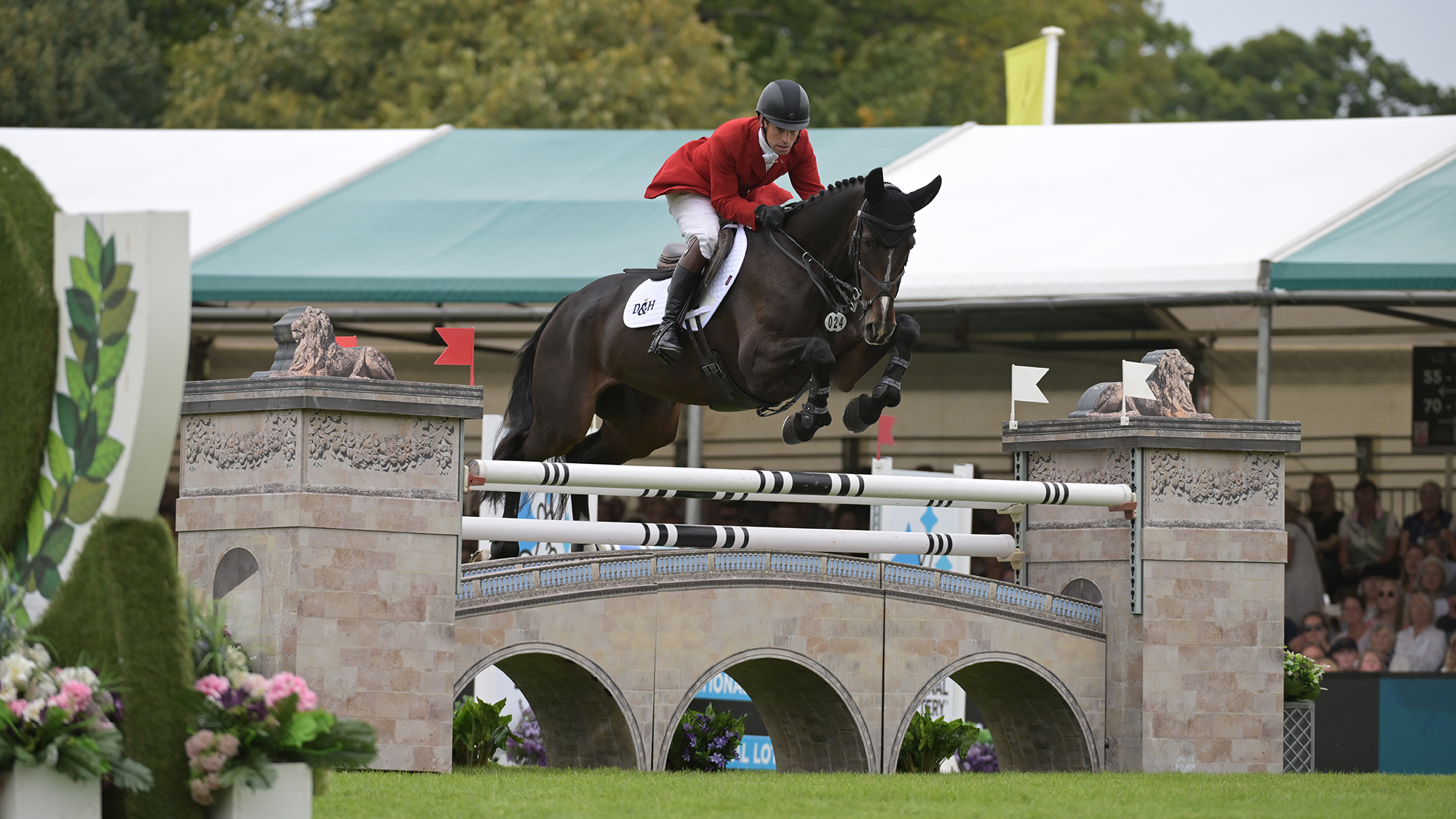 Harry Meade, a rider in a red coat, rides dark bay horse Cavalier Crystal over a wall fence in the showjumping at Burghley 2025