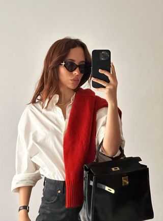 Female fashion influencer Debora Rosa poses for a mirror selfie wearing black cat eye sunglasses, a white button-down shirt, a red sweater draped on the shoulder, Hermes bag, and black jeans