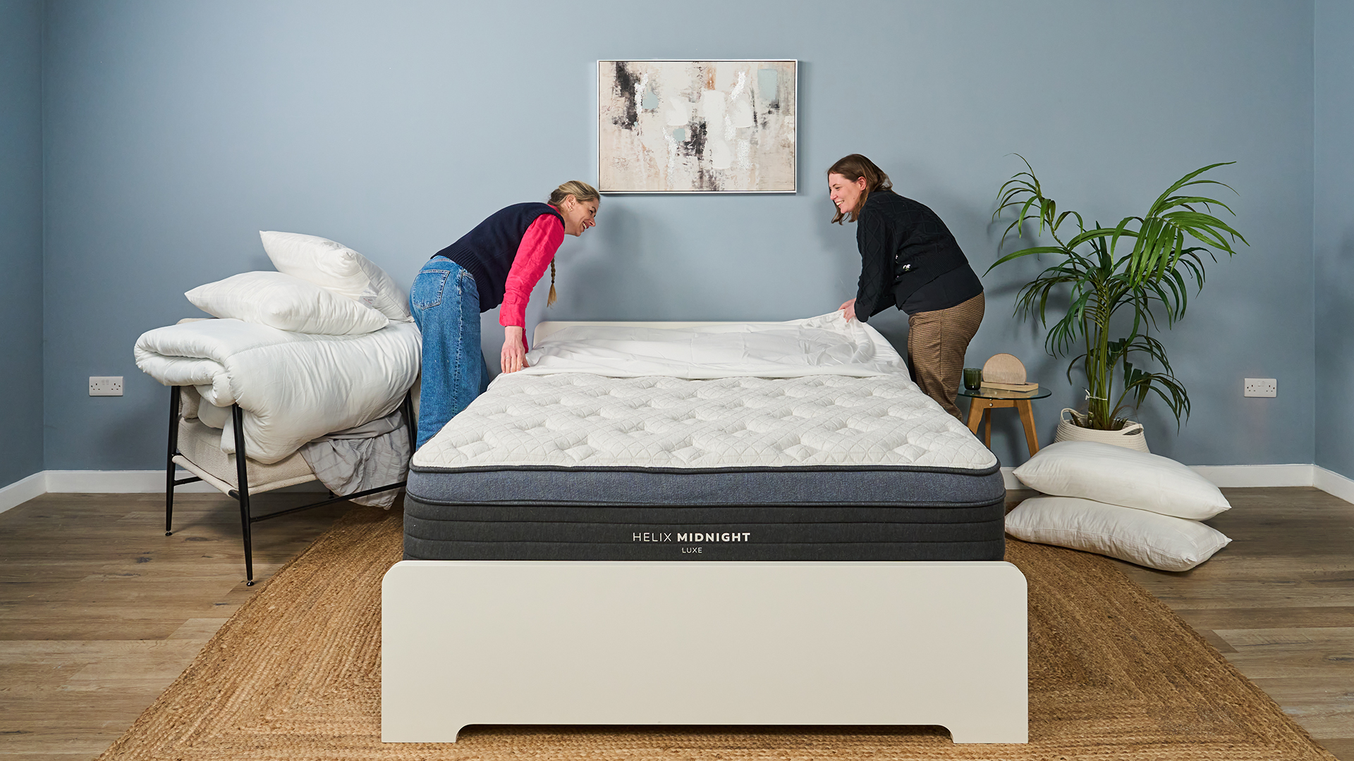 Two women remove the bed sheets from a Helix Midnight Luxe mattress, leaving the bed bare.