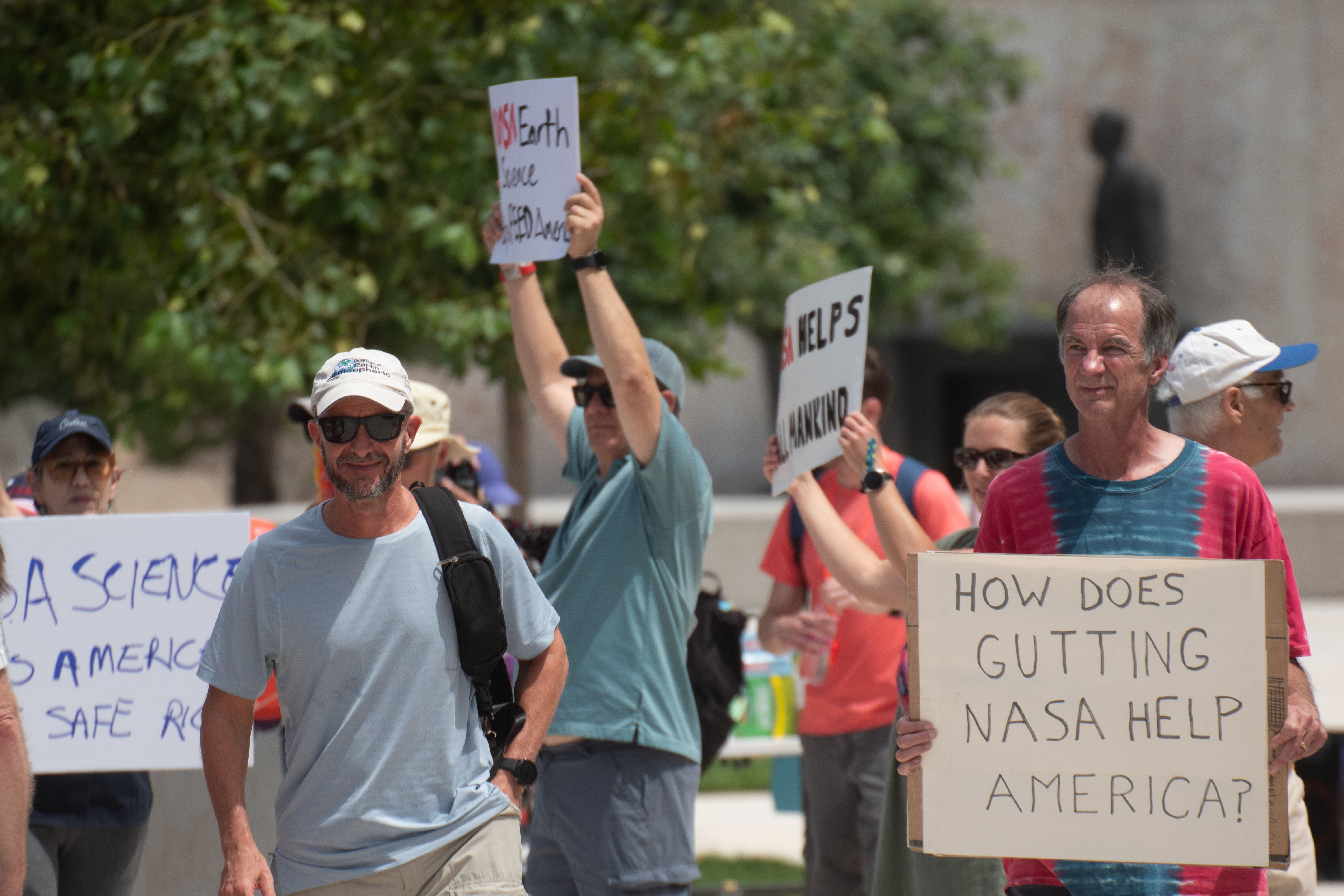 People hold signs outside on a sunny day in protest of NASA budget cuts, July 20, 2025.