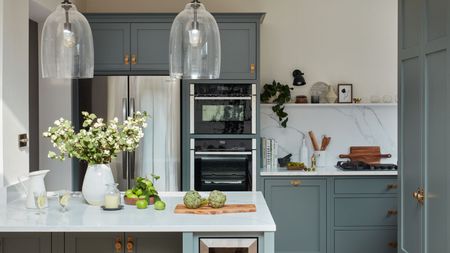 A teal blue and white kitchen with stainless steel appliances and white marble backsplashes. There is an island and overhead lighting visible in foreground