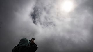a mostly cloudy sky obscures the sun and a person is below holding a camera up towards the sky.