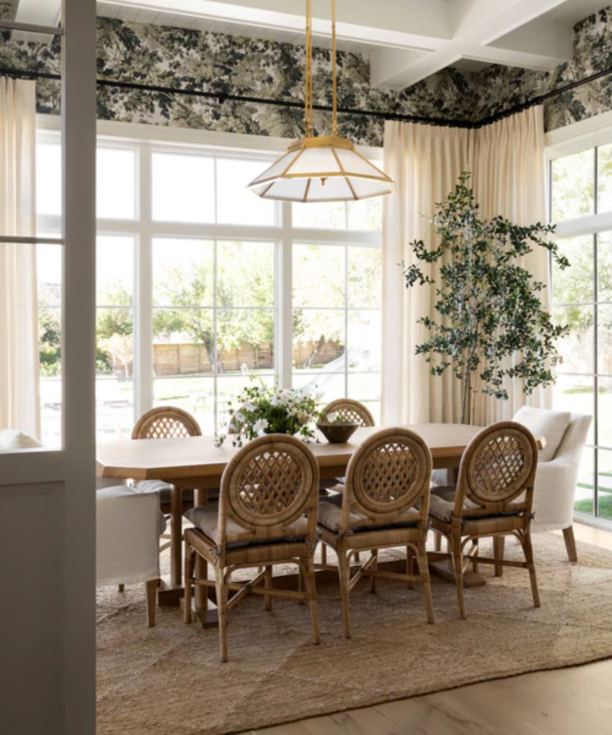 light and bright dining room with large windows and green leafy wallpaper with a jute rug, wooden table, wood and rattan chairs, and two linen armchairs