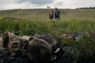 A closeup of human skeletons in a field with (L to R) Bella Ramsey as Ellie and Pedro Pascal as Joel in the background in The Last of Us episode 3 on HBO