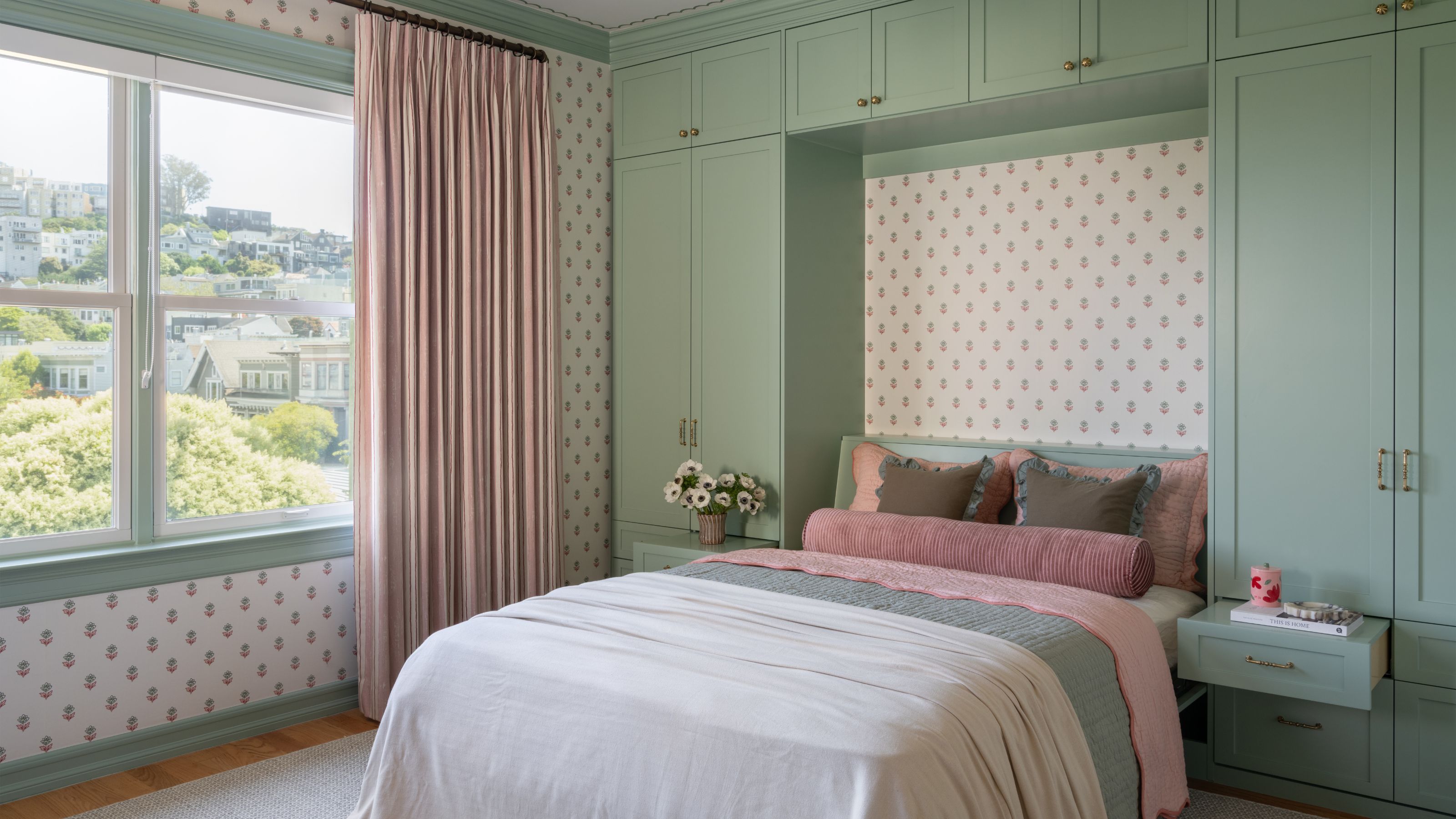 A bedroom featuring a bed built into an alcove framed by sage green cabinetry, with pink and green patterned wallpaper and a large window with striped pink curtains