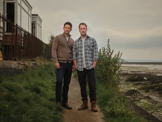 Matthew Venn (Ben Aldridge) with his husband Jonathan (Declan Bennett) standing on the path outside their house, with the beach in the background