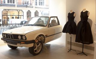 A display in a store with two sleeveless black dresses on mannequins one short and one long next to an old white car with bouquets of flowers inside of it.