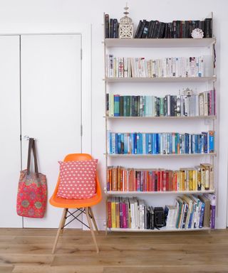 A white room with a tall bookshelf with black, white, green, blue, red, and multicolored books cascading down it with an orange curved chair with a red throw pillow on it with a red and black bag on the door next to it
