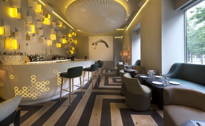 A bar with a white curved counter, decorated tables, grey sofa's, grey chairs, light and dark wood flooring and yellow wall lights.
