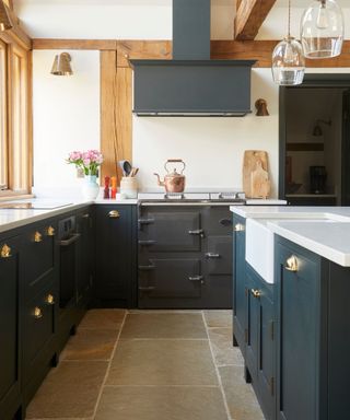 A kitchen with dark blue cabinets with gold cabinet pulls, a black oven with a copper kettle on the stove and a dark hood above it, and light wooden beams on the white painted walls and two glass pendant lights hanging