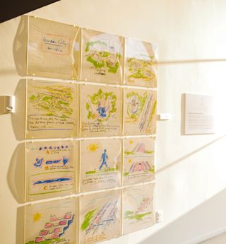 Coloured sketches on yellow paper hang on the wall in a grid