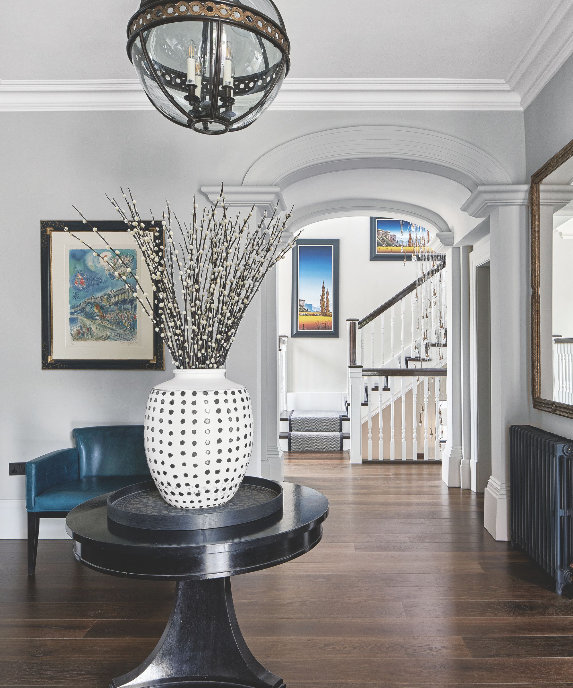 A bright entryway with wooden floors, white walls, a black circular table with a white vase and dried flowers, a blue leather chair, artwork, a painted radiator and gold framed mirror. Behind, a staircase is seen with bright and colorful framed artwork.