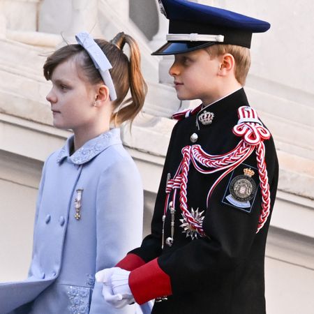 Prince Jacques wearing a military uniform and Princess Gabriella wearing a lilac dress coat and headband walking outside the palace in Monaco