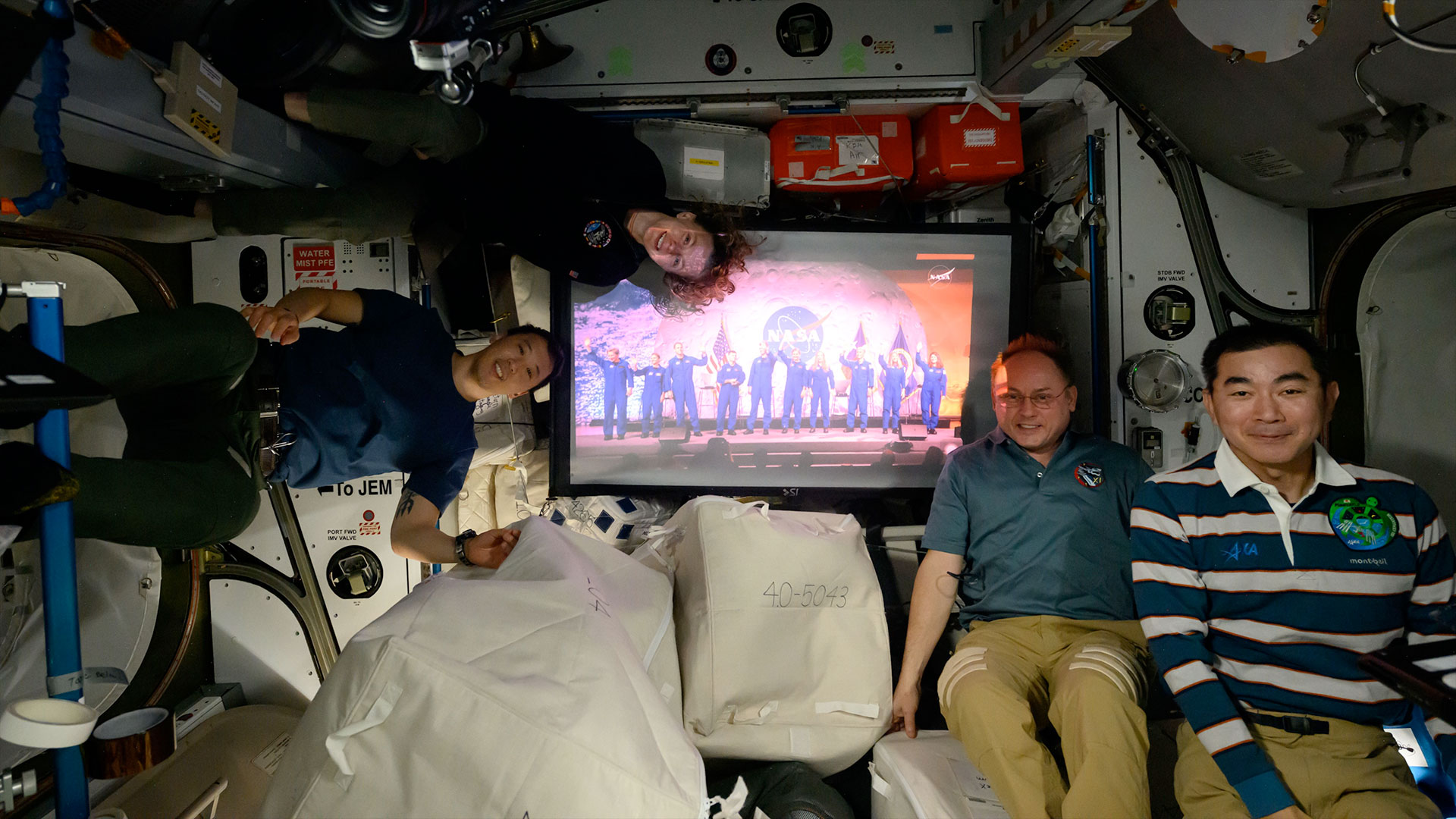 three men and a woman in polo shirts and slacks float on a space station near a screen showing 10 people in blue flights.