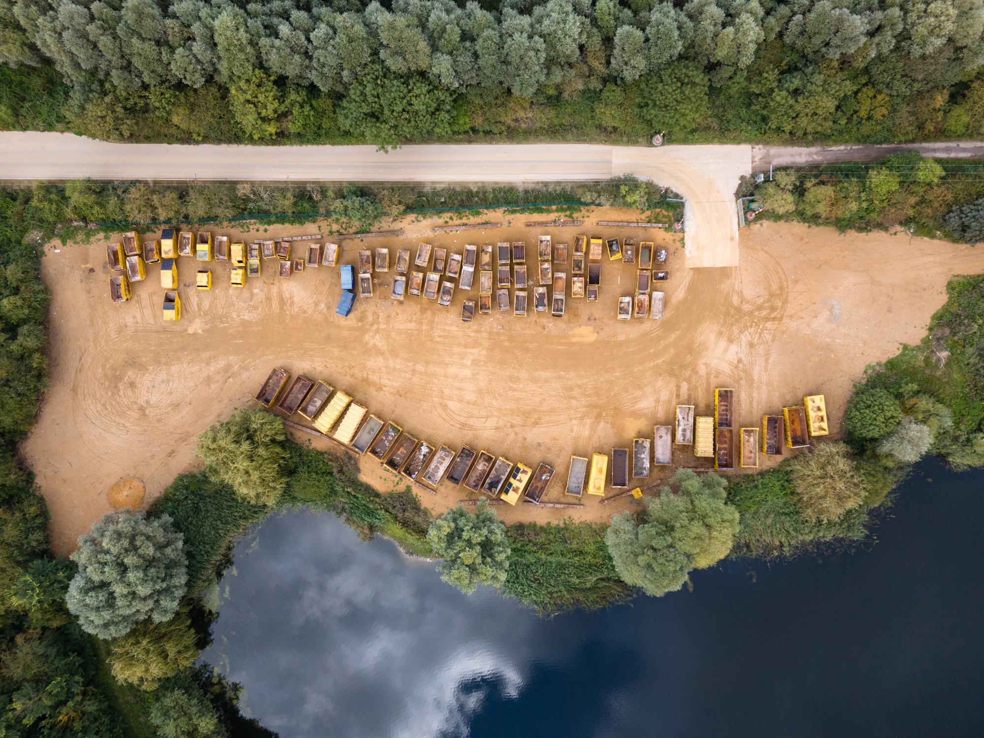 aerial view of a dirt lot below an adjacent road, neighbored by a thick row of tress above. Below the lot sits a body of water.  various yellow containers are parcked.
