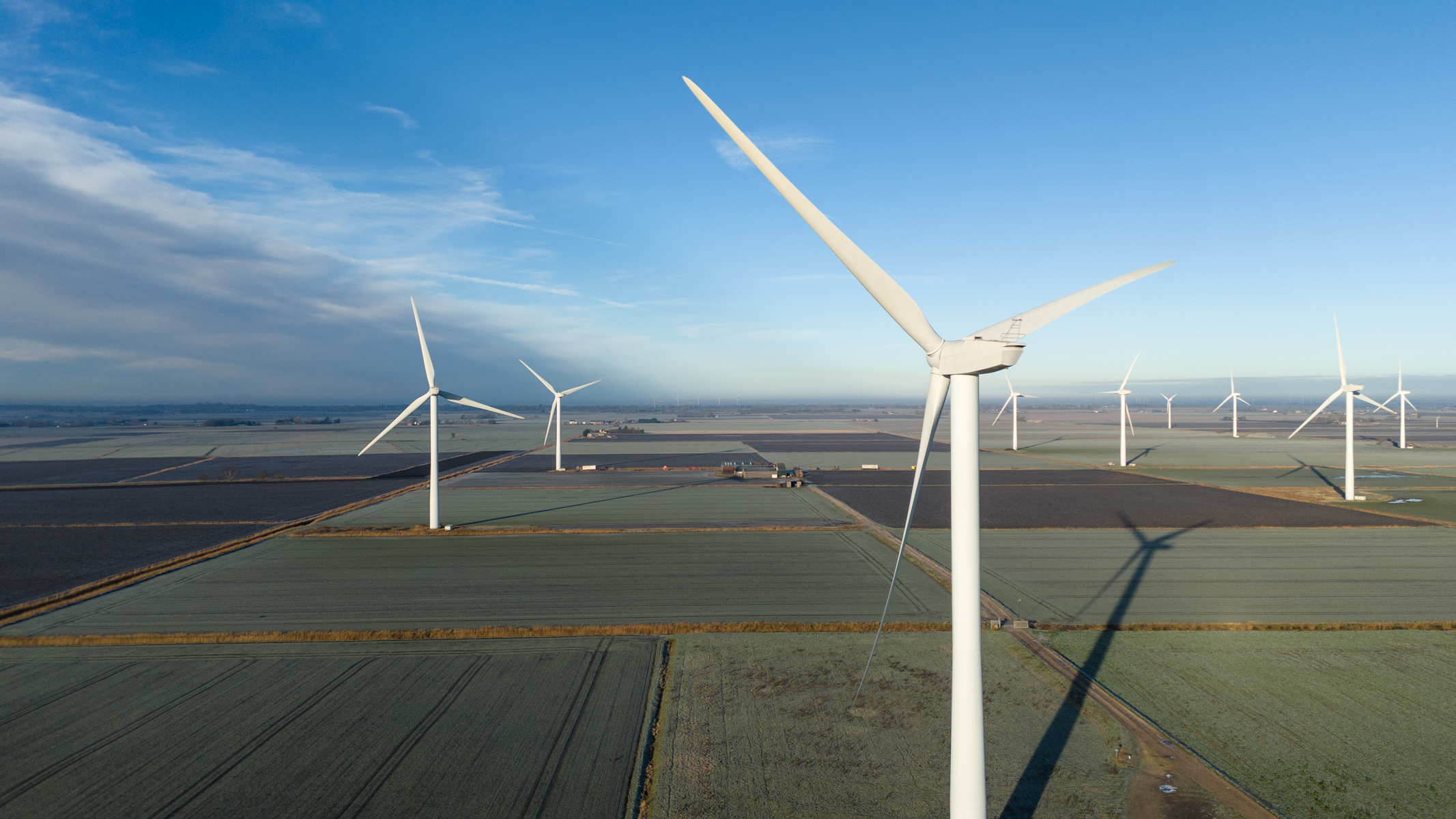 Some wind turbines in open green fields with blue skies in the centre and right of the photo with gray clouds to the left.