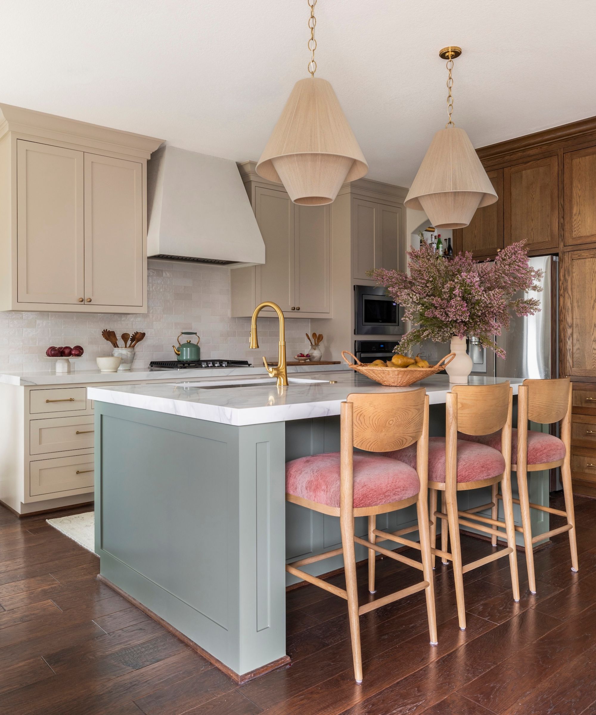 A modern kitchen with beige cabinets, dark wood flooring, and a sage green kitchen island with a white marble countertop. Three pink bar chairs, brass fixtures, and two modern beige pendant lights