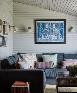 Living room with white paneled walls and ceiling, blue corner sofa, artwork and framed images on wall, four matching wall lamps with shades, dark wood coffee table