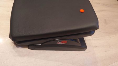 Footrest with a curved black cushion on wood flooring. An orange-colored button on the top adds contrast. The design suggests ergonomic comfort.
