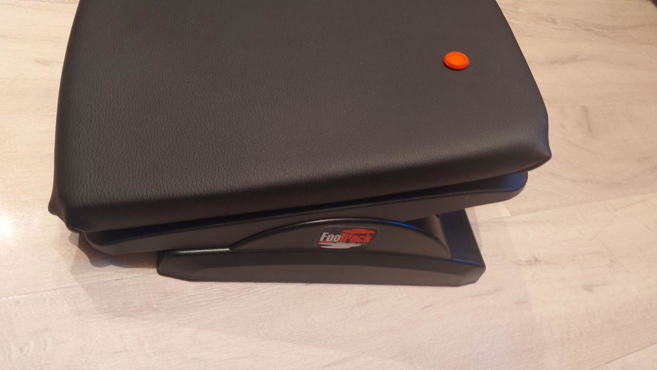 Footrest with a curved black cushion on wood flooring. An orange-colored button on the top adds contrast. The design suggests ergonomic comfort.